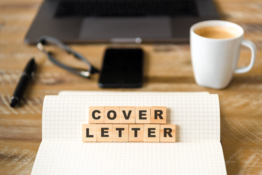 Closeup on notebook over wood table background, focus on wooden blocks with letters making COVER LETTER words. Business concept image. Laptop, glasses, pen and mobile phone in a defocused background.